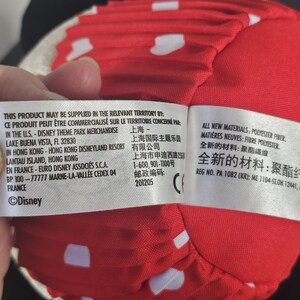 May include: A red plush Minnie Mouse bow with white polka dots. The bow is made of a soft material and is part of a Disney theme park merchandise collection. The bow is a classic accessory.