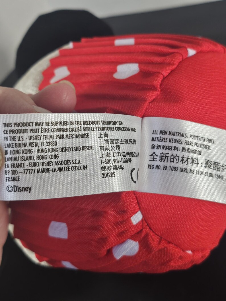 May include: A red plush Minnie Mouse bow with white polka dots. The bow is made of a soft material and is part of a Disney theme park merchandise collection. The bow is a classic accessory.