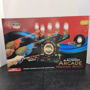 May include: A red, black, and blue electronic arcade hover shot game with two toy guns. The game features a target with five holes and a digital display. The box includes the text "2 Player Electronic Arcade Hover Shot" and "Special Edition 2 Player Shoot-Out!"