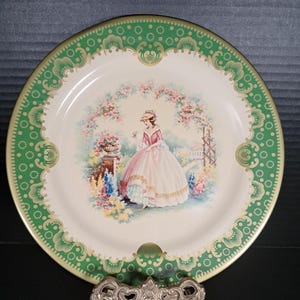 May include: Decorative plate with a green and gold rim featuring a Victorian-era woman in a pink dress amidst a floral garden scene. The plate is displayed on a silver-colored stand, showcasing a vintage aesthetic.