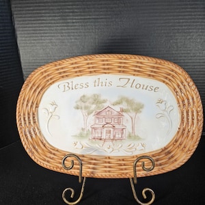 May include: Oval decorative plate with a woven border and a central image of a house and trees. The plate has the words "Bless this House" in gold lettering. The plate is displayed on a gold metal stand.