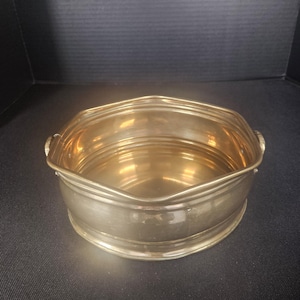 May include: A silver-colored, decorative bowl with a scalloped rim and two small handles. The bowl has a round base and a series of horizontal ridges around the body. The interior of the bowl reflects a golden hue.