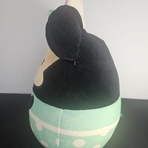 May include: A plush toy shaped like a cartoon character, featuring a black head with large ears and a light-colored face. The body is a light blue with white polka dots and a white stripe. The toy has a pink bow and a green bow.