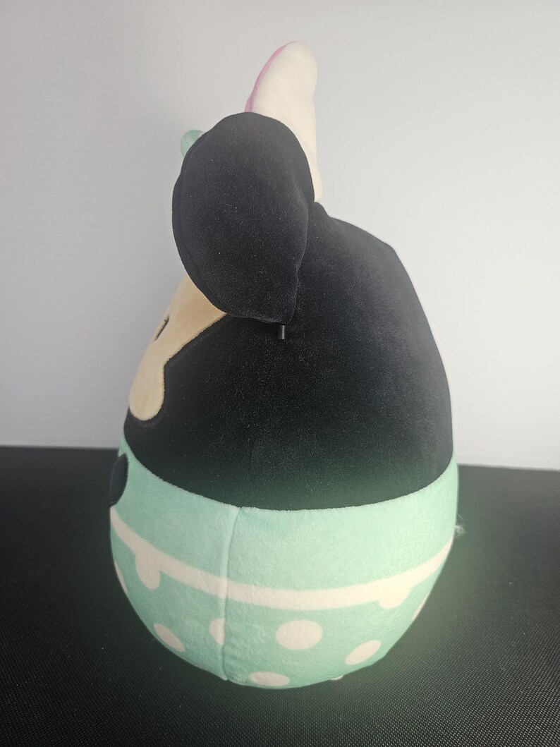 May include: A plush toy shaped like a cartoon character, featuring a black head with large ears and a light-colored face. The body is a light blue with white polka dots and a white stripe. The toy has a pink bow and a green bow.
