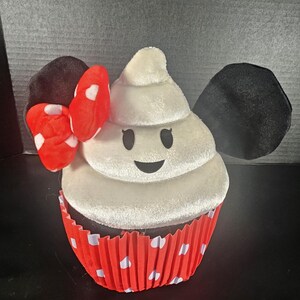 May include: A plush cupcake toy with a white frosting design, black eyes, and a smiling mouth. The cupcake sits in a red and white polka dot liner. Black ears and a red bow with white polka dots complete the design.