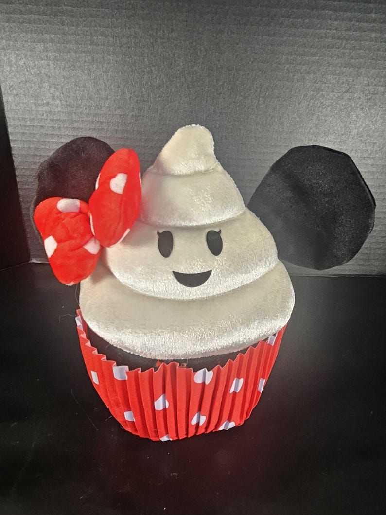 May include: A plush cupcake toy with a white frosting design, black eyes, and a smiling mouth. The cupcake sits in a red and white polka dot liner. Black ears and a red bow with white polka dots complete the design.