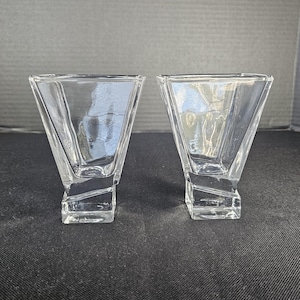 May include: Two clear glass vessels featuring a wide, flared top opening that tapers down into a thick, angular, and faceted base. The design incorporates geometric lines and a heavy, solid glass bottom, providing a stable foundation for each piece.