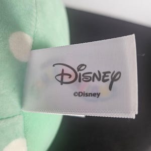 May include: A close-up of a plush toy with a mint green body and white polka dots. A white tag with the Disney logo and the text "&copy;Disney" is attached to the toy.