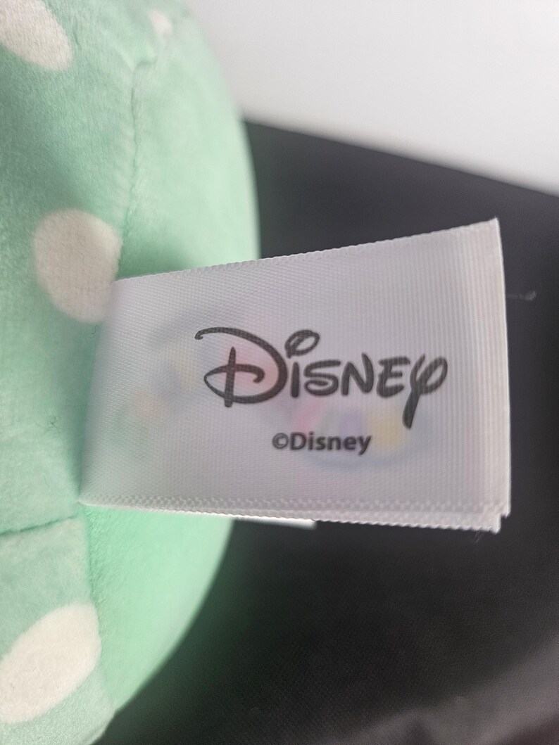 May include: A close-up of a plush toy with a mint green body and white polka dots. A white tag with the Disney logo and the text "&copy;Disney" is attached to the toy.