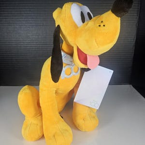 May include: A plush Pluto dog toy in bright yellow with black ears and nose, and white eyes. The dog has a pink tongue sticking out and a light blue collar. A white tag with the Disney 100 logo is attached.