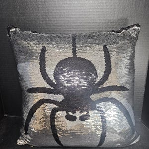 May include: A square decorative pillow with reversible sequins. The pillow features a large black spider design on a silver and black background. The sequins change color when brushed, creating a shimmering effect. The pillow is a Halloween or spooky-themed home decor item.
