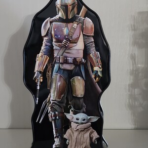 Puede incluir: Una fiambrera de metal negro con un personaje de la serie Star Wars, el Mandaloriano. El Mandaloriano lleva un casco y una armadura y sostiene un arma. Un peque&ntilde;o personaje verde, Grogu, est&aacute; de pie frente al Mandaloriano.