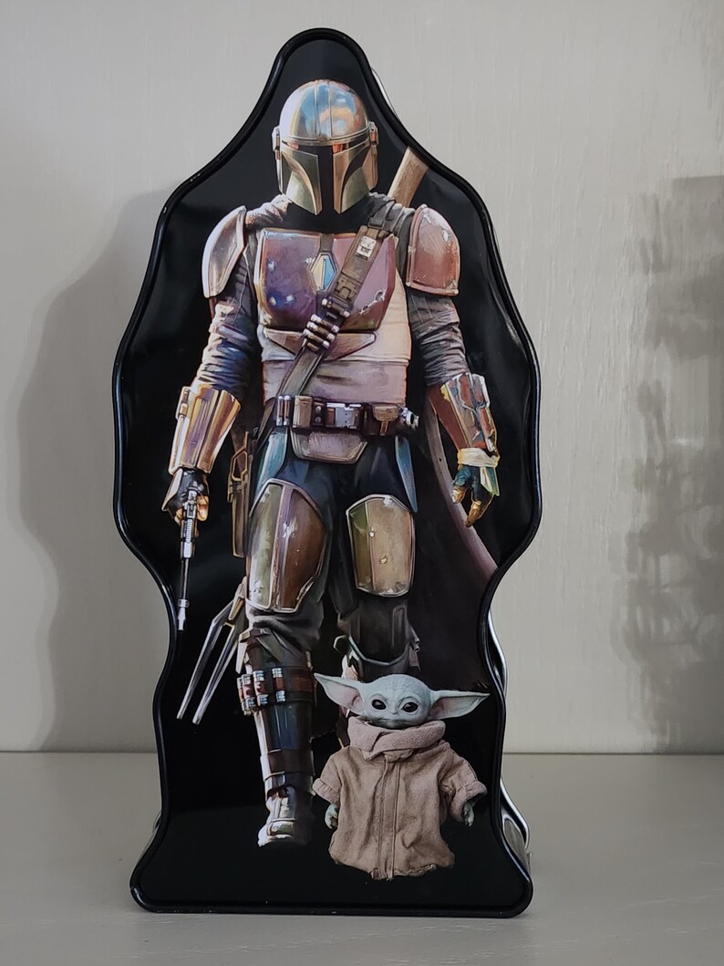 Puede incluir: Una fiambrera de metal negro con un personaje de la serie Star Wars, el Mandaloriano. El Mandaloriano lleva un casco y una armadura y sostiene un arma. Un peque&ntilde;o personaje verde, Grogu, est&aacute; de pie frente al Mandaloriano.
