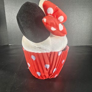 May include: A plush cupcake toy featuring a black ear, a red bow with white polka dots, and a white frosting top. The cupcake is in a red paper cup with white polka dots. A fun, whimsical toy.