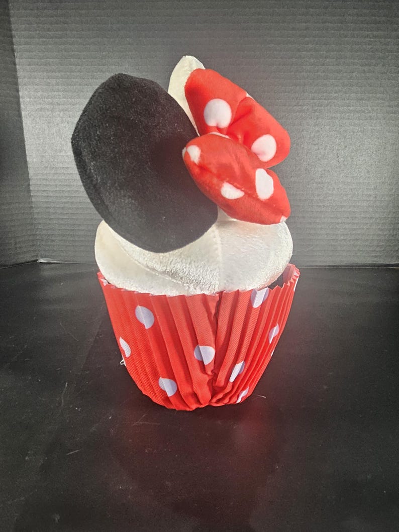 May include: A plush cupcake toy featuring a black ear, a red bow with white polka dots, and a white frosting top. The cupcake is in a red paper cup with white polka dots. A fun, whimsical toy.