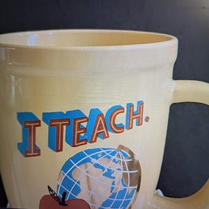 May include: A cream-colored ceramic mug featuring the text I TEACH. in blue and orange block letters. Below the text is a graphic of a blue and white globe and a red apple with a black stem. The mug has a smooth finish and a curved handle.