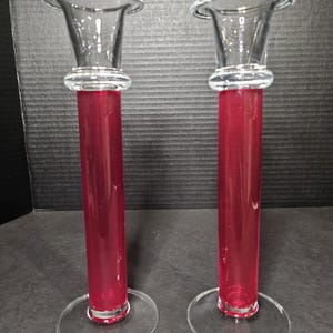 May include: A set of two clear glass candle holders with a red base. The holders have a flared top and a round base.