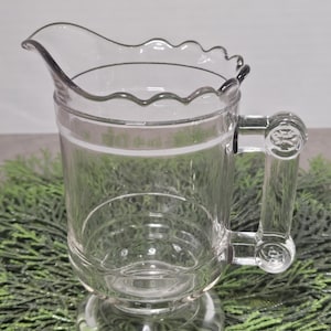 May include: A clear glass pitcher with a scalloped rim and a single white band around the middle. The pitcher has a handle with a decorative knob and a pedestal base.
