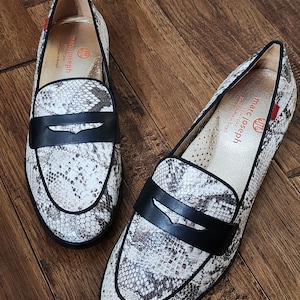 May include: A pair of snakeskin loafers with black leather accents. The loafers have a pointed toe and a low heel. The brand name "marc joseph new york" is visible on the inside of the shoes.
