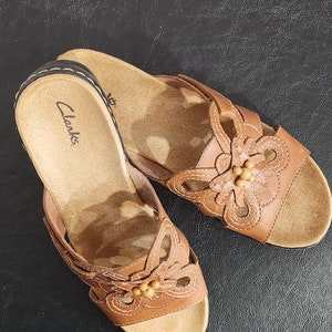 May include: A pair of tan leather slide sandals featuring a floral cutout design on the strap with small bead accents in the center. The footbed is tan with the Clarks brand name printed in black near the heel and a dark brown stitched edge.