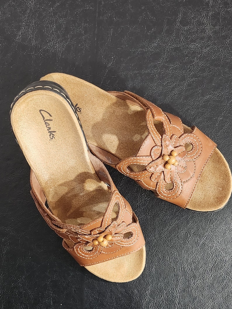 May include: A pair of tan leather slide sandals featuring a floral cutout design on the strap with small bead accents in the center. The footbed is tan with the Clarks brand name printed in black near the heel and a dark brown stitched edge.