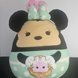 May include: A plush toy shaped like Minnie Mouse, featuring black ears, a light yellow face, and a mint green body with white polka dots. The toy has a pink and mint green bow and a basket with a pink bunny.