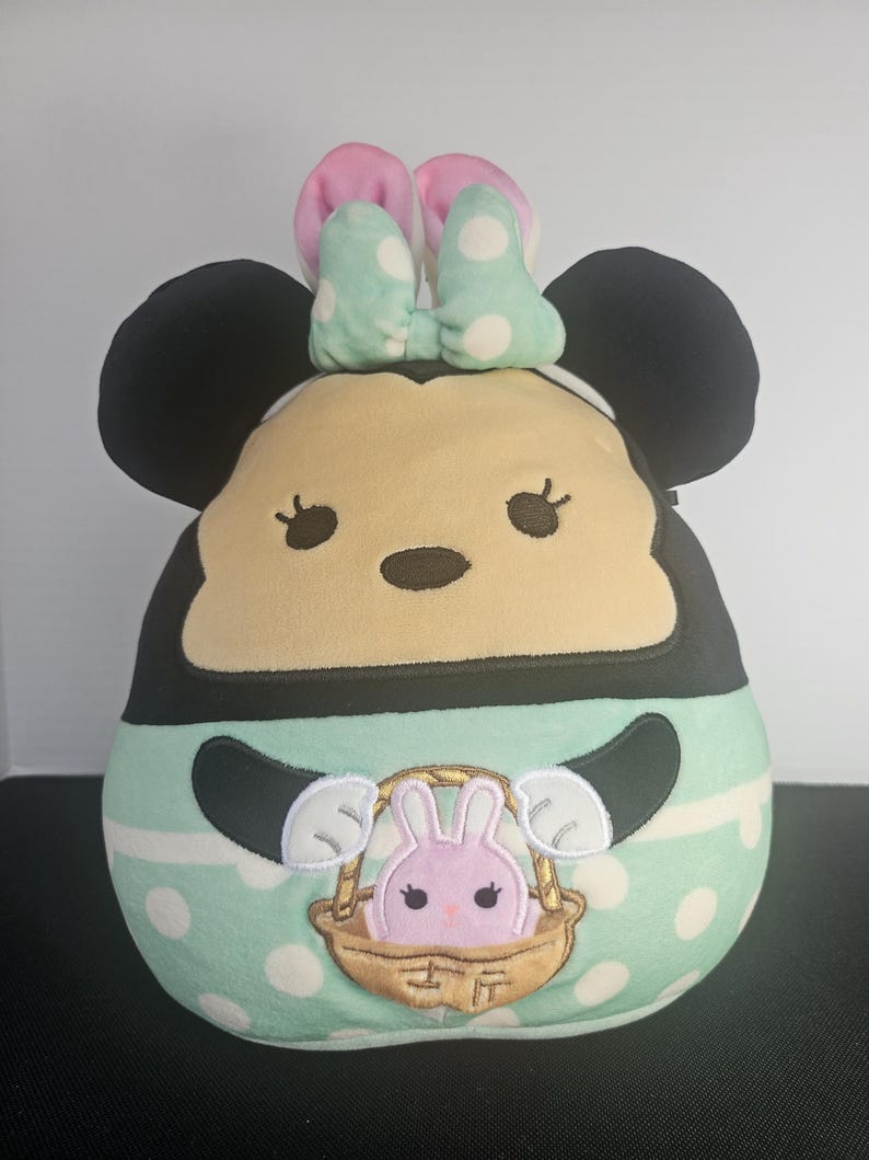 May include: A plush toy shaped like Minnie Mouse, featuring black ears, a light yellow face, and a mint green body with white polka dots. The toy has a pink and mint green bow and a basket with a pink bunny.