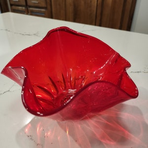 May include: A red glass bowl with a wavy, ruffled rim. The bowl is made of clear glass with a red tint.
