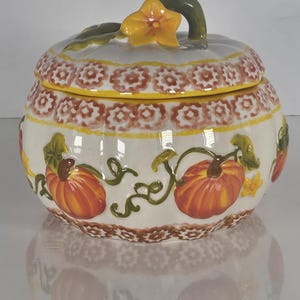 May include: A ceramic pumpkin-shaped jar with a lid. The jar is white with orange pumpkins and green vines painted on the sides. The lid has a yellow flower and green stem handle. The rim has a brown and white pattern.