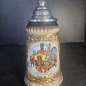 May include: A ceramic beer stein with a metallic lid featuring a relief-molded scene of a horse-drawn carriage and the Frauenkirche in Munich. The central graphic includes the text Oktoberfest München and gold-painted decorative accents.