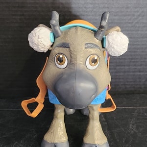 Pu&ograve; includere: Un giocattolo di plastica a forma di renna con grandi occhi marroni, corna grigie e un naso grigio. La renna ha paraorecchie bianche, un'imbracatura arancione e un collare blu e viola. Il giocattolo &egrave; in piedi su quattro zampe.