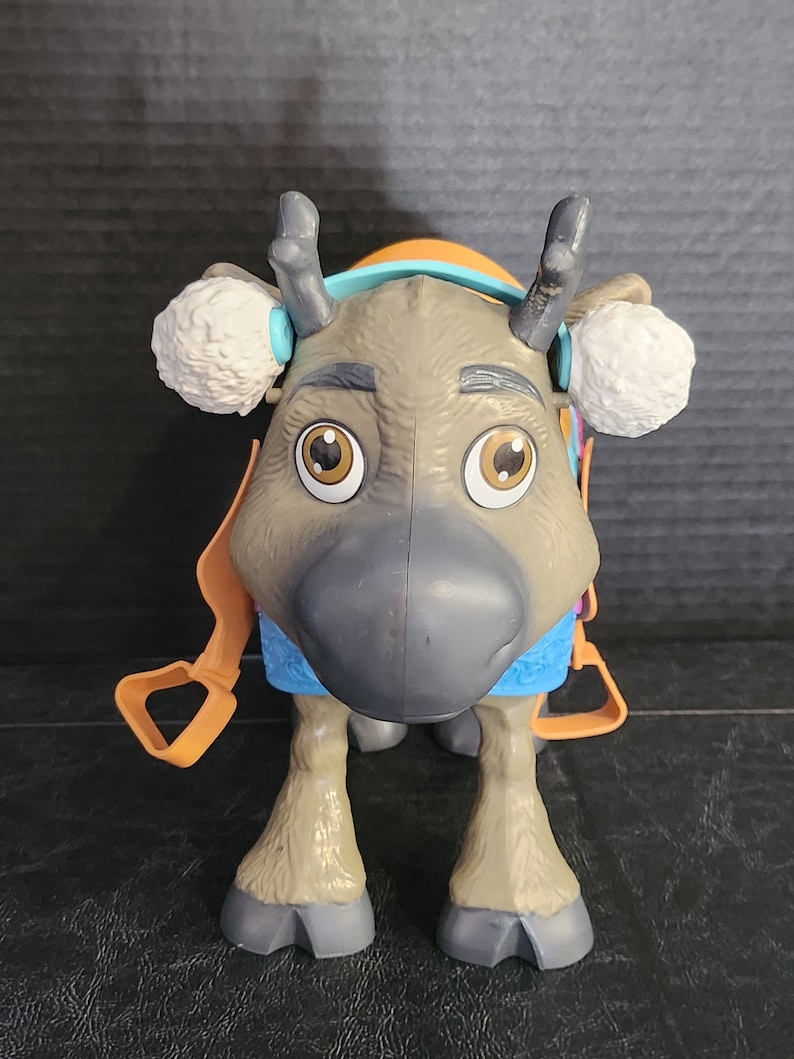 Pu&ograve; includere: Un giocattolo di plastica a forma di renna con grandi occhi marroni, corna grigie e un naso grigio. La renna ha paraorecchie bianche, un'imbracatura arancione e un collare blu e viola. Il giocattolo &egrave; in piedi su quattro zampe.