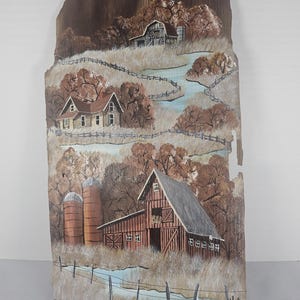 May include: Hand-painted wooden wall art depicting a rural landscape. The artwork features a barn, silos, houses, and a winding river. The color palette includes shades of brown, beige, and blue, creating a rustic and serene scene. The artwork is mounted on a wooden plank.