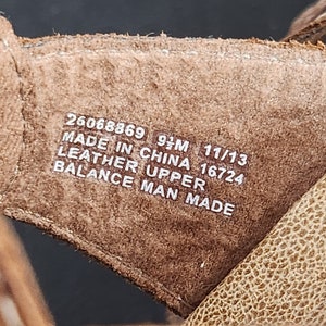 May include: A brown leather shoe interior featuring white printed text that reads 26068869 9 1/2M 11/13 MADE IN CHINA 16724 LEATHER UPPER BALANCE MAN MADE. The material has a textured surface and visible stitching along the edges of the brown leather strap.