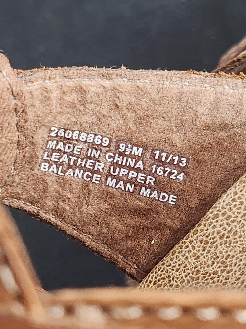 May include: A brown leather shoe interior featuring white printed text that reads 26068869 9 1/2M 11/13 MADE IN CHINA 16724 LEATHER UPPER BALANCE MAN MADE. The material has a textured surface and visible stitching along the edges of the brown leather strap.