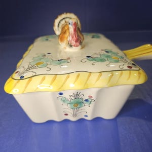 May include: A white ceramic casserole dish with a yellow and blue floral design. The lid features a small ceramic turkey figurine.