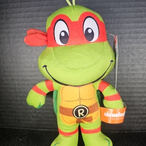 May include: A plush Teenage Mutant Ninja Turtle toy in green, red, and orange. The toy has a red mask, orange stripes on the arms and legs, and a brown belt with the letter "R". A small orange tag with the Nickelodeon logo is attached.