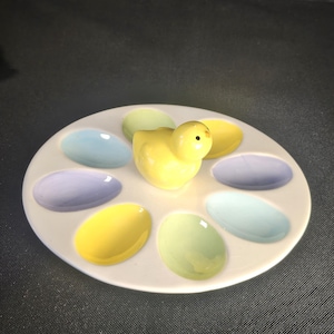 May include: A white ceramic egg holder with eight oval-shaped compartments in pastel colors. A yellow chick figurine sits in the center. The egg holder is a decorative item, perfect for Easter.