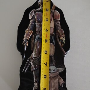 Puede incluir: Una fiambrera de metal negro con un gr&aacute;fico del Mandaloriano y Grogu de la serie Star Wars. El Mandaloriano lleva su armadura caracter&iacute;stica y sostiene un rifle bl&aacute;ster. Grogu est&aacute; de pie frente a &eacute;l, mirando al espectador. La fiambrera mide aproximadamente 23 cm de alto.