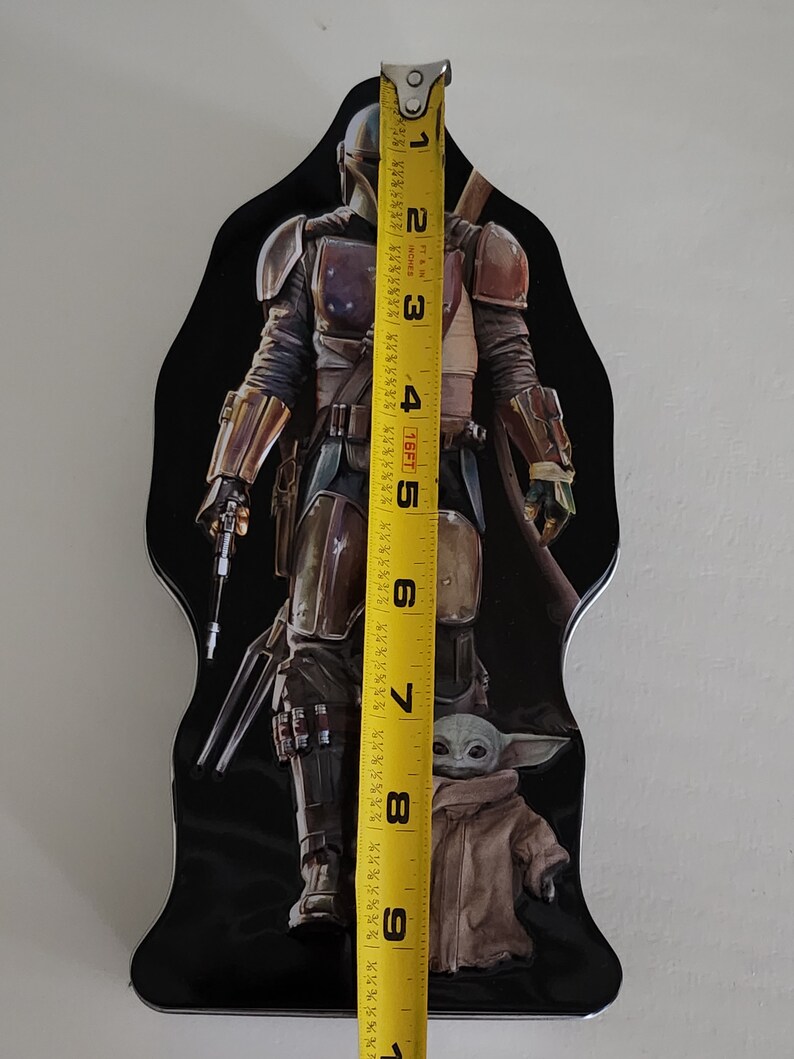 Puede incluir: Una fiambrera de metal negro con un gr&aacute;fico del Mandaloriano y Grogu de la serie Star Wars. El Mandaloriano lleva su armadura caracter&iacute;stica y sostiene un rifle bl&aacute;ster. Grogu est&aacute; de pie frente a &eacute;l, mirando al espectador. La fiambrera mide aproximadamente 23 cm de alto.