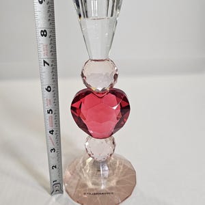 Op de afbeelding: Een heldere kristallen kandelaar met een facetontwerp. De kandelaar heeft een rood hartvormig kristal in het midden, met heldere kristallen bollen en een voet. De hoogte is ongeveer 20 cm.