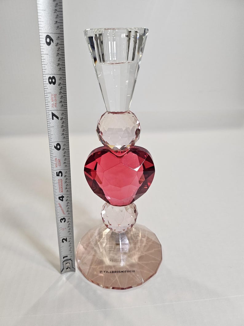 Op de afbeelding: Een heldere kristallen kandelaar met een facetontwerp. De kandelaar heeft een rood hartvormig kristal in het midden, met heldere kristallen bollen en een voet. De hoogte is ongeveer 20 cm.