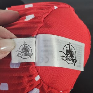May include: Close-up of a red fabric with white polka dots. The fabric is part of a larger object, possibly a hat or accessory. Two Disney tags are attached, featuring the Disney logo.