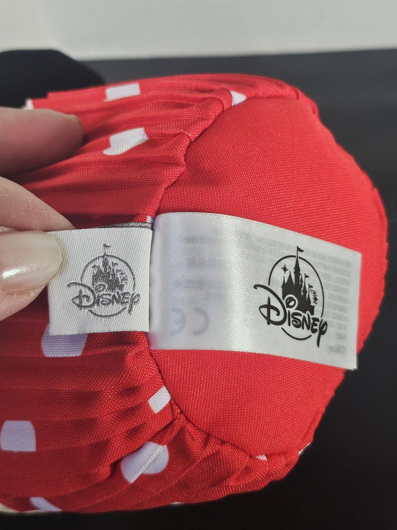 May include: Close-up of a red fabric with white polka dots. The fabric is part of a larger object, possibly a hat or accessory. Two Disney tags are attached, featuring the Disney logo.