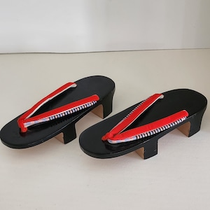 May include: A pair of black wooden geta sandals with red and white striped straps. The sandals have a raised wooden platform.