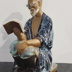May include: Ceramic figurine of a man wearing glasses and a blue and white robe, sitting with a book in his hands.