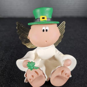 May include: A ceramic figurine of an angel wearing a green leprechaun hat with a gold buckle. The angel has gold wings, a white robe, and is holding a green shamrock. The figurine has a light skin tone and is sitting down.