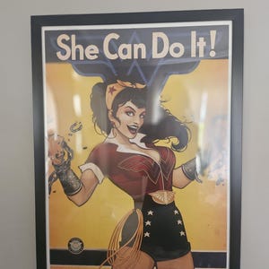 May include: Framed poster featuring a vintage-style illustration of a woman in a Wonder Woman-inspired outfit. The poster has the text "She Can Do It!" at the top. The woman is smiling and breaking chains, with a yellow and red color scheme.