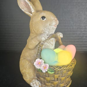 May include: A tan-colored bunny figurine holding a woven basket filled with pastel-colored Easter eggs. The bunny has long ears and is decorated with pink flowers. The basket is brown with a rope handle.
