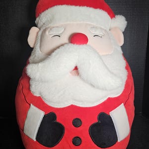 May include: A plush Santa Claus pillow in a seated position. The pillow features a red hat and suit with white trim, a white beard and mustache, a red nose, and closed eyes. Black gloves and white patches are on the front.